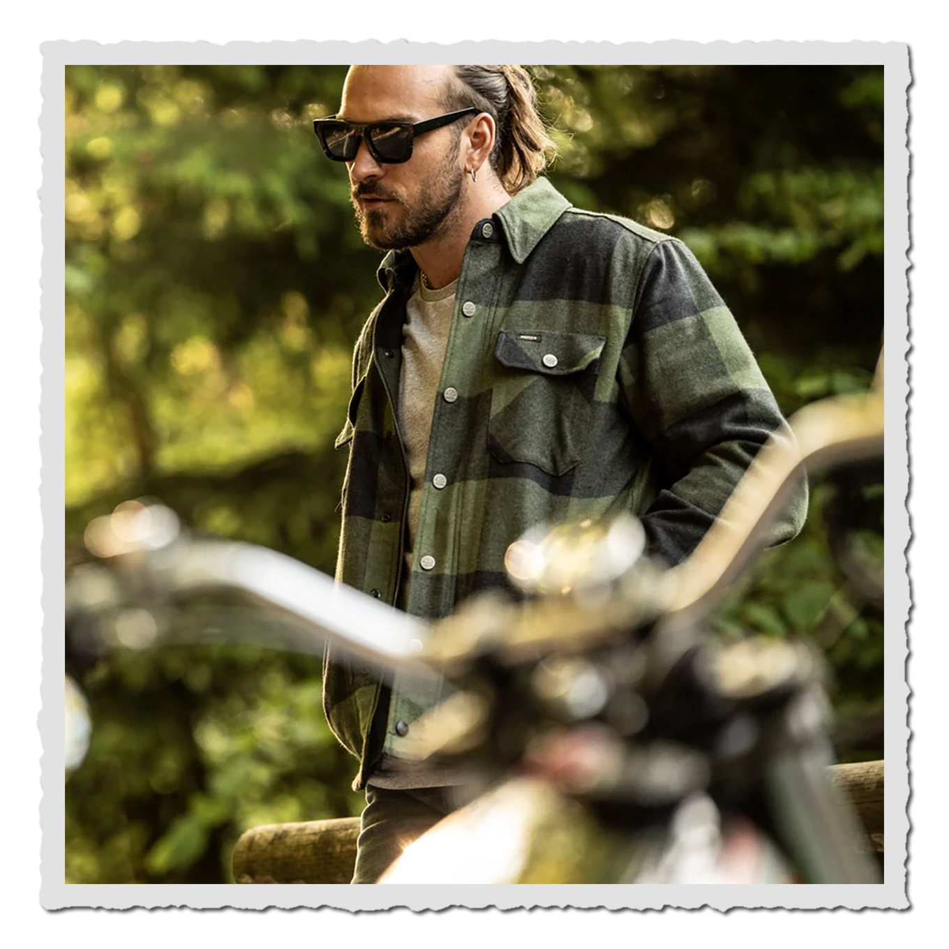 Person mit Sonnenbrille im karierten Rider Shirt, Seitenansicht im Wald, Motorrad unscharf im Vordergrund.